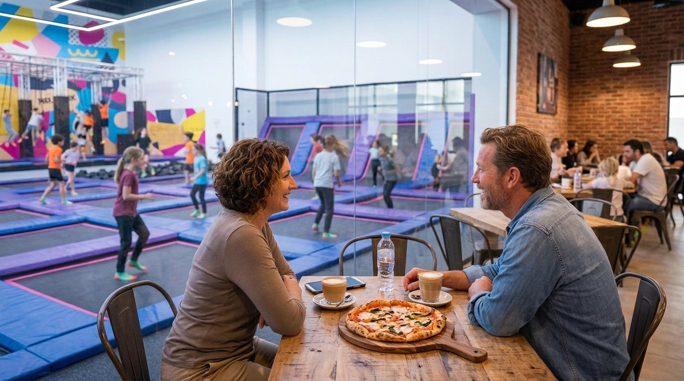 Parents relaxing at a cafe table while children jump on trampolines in the background